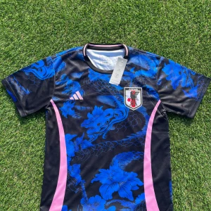 Japan Adidas blå/svart fotbollströja L - Japan landslagströja från Adidas i slim fit. Svart bas med blått drak- och blommönster, rosa Adidas-logga och rosa detaljer längs sidorna. Broderad Japan-emblem på bröstet. Tillverkad i lätt och ventilerande material, perfekt för match eller träning.