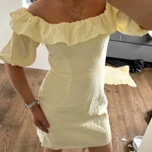 Gul offshoulder kortklänning med volang - Superfin ljusgul kortklänning med offshoulder och volang runt hela överdelen. Klänningen har puffiga korta ärmar och en figurnära passform. Perfekt för sommarens alla tillfällen och ger en riktigt fräsch vibe.