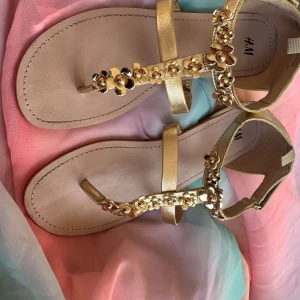 Guldiga sandaler med blommor från H&M - Snygga sandaler i guld från H&M med blommiga detaljer på remmarna. Platt sula och öppen tå, perfekt för varma dagar.