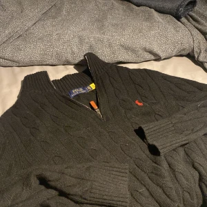 Svart stickad kofta Polo Ralph Lauren - Svart kabelstickad kofta från Polo Ralph Lauren med halv dragkedja och klassisk röd logga broderad på bröstet. Tröjan har ribbade muddar och hög krage, perfekt för lager-på-lager. Materialet är mjukt och värmande ull.