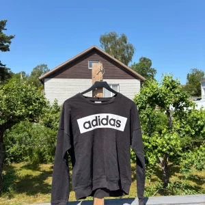 Svart Adidas sweatshirt med logga - Svart sweatshirt från Adidas med stor vit logga framtill. Tröjan har rund halsringning och långa ärmar. Tillverkad i mjuk bomullsmix, perfekt för en sportig och avslappnad stil. 