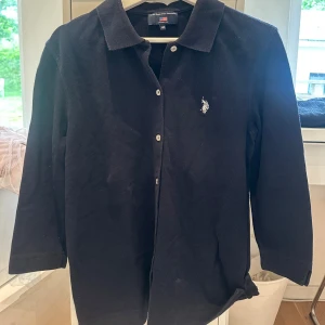 Svart långärmad skjorta  - Klassisk svart skjorta från U.S. Polo Assn. med vit broderad logga på bröstet. Skjortan har lång ärm, knappar framtill och en rak passform. 