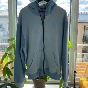 Ljusblå hoodie från Verborgen Studios  - Säljer en ljusblå hoodie från Verborgen Studios med glittrande text framtill. Tröjan har dragkedja, huva och stora fickor på magen. Perfekt för dig som gillar en chill och oversized look. Materialet känns mjukt och skönt, troligen bomull eller polyester.