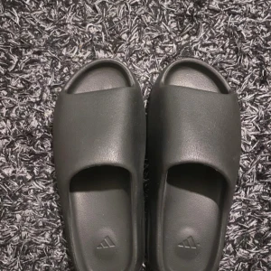 Adidas svarta slides med räfflad sula - Svarta Adidas slides med minimalistisk design och öppen tå. Tillverkade i syntetmaterial med en chunky, räfflad sula som ger cool vibe. Perfekta för chill dagar eller när du vill ha något bekvämt och stilrent på fötterna.