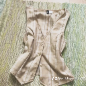 Beige rutig väst från H&M Divided - Snygg beige väst med rutigt mönster från H&M Divided. Västen har v-ringning, knappar framtill och en figurnära passform. Perfekt att styla över en t-shirt eller skjorta för en trendig look.