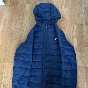 Blå pufferjacka från Lyle & Scott - Snygg marinblå pufferjacka från Lyle & Scott med huva och dragkedja. Jackan har det klassiska gula fågel-loggan på bröstet och är quiltad för extra värme. Perfekt för kyliga dagar och enkel att matcha med det mesta.