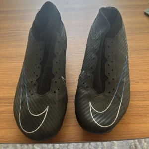 Nike Air Zoom Mercurial fotbollsskor - Svarta Nike Air Zoom Mercurial fotbollsskor med vita och blå detaljer. Skorna har en låg profil, rund tå och mönstrad ovandel i syntetmaterial. Sulan har fasta dobbar för bra grepp på planen. Snygg design med stora Swoosh-loggor på tån och sidan.