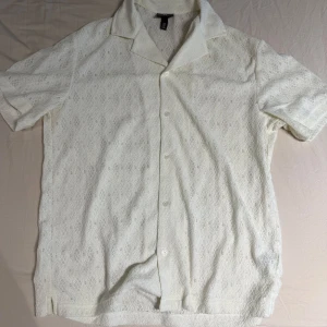 Vit spetsmönstrad skjorta H&M - Vit kortärmad skjorta från H&M med spetsliknande mönster och klassisk krage. Skjortan har knäppning framtill och är tillverkad i ett lätt material som passar perfekt för varma dagar. Snygg och luftig med unik struktur.