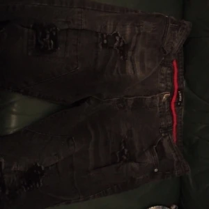 Svarta slitna jeans med röd detalj från Encrypted - Säljer ett par svarta jeans från Encrypted med coola slitningar och en röd detalj i midjan. Jeansen har klassisk femficksdesign och raka ben. Perfekta för dig som gillar snygga jeans 