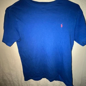 Blå t-shirt från Polo Ralph Lauren - Klassisk blå t-shirt från Polo Ralph Lauren med korta ärmar och rund hals. T-shirten har den ikoniska lilla rosa loggan broderad på bröstet. Tillverkad i mjuk bomull som känns skön mot huden. Perfekt för en clean och stilren look.