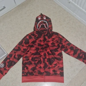 BAPE röd camo shark hoodie - Säljer en ikonisk BAPE shark hoodie i röd camo med dragkedja hela vägen upp i huvan. Huvan har hajmotiv med tänder och ögon, samt broderade bokstäver och tigerdetalj. Jackan har fickor fram och är perfekt för dig som vill sticka ut.