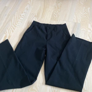 Svarta kostymbyxor från Grunt - Svarta kostymbyxor från Grunt med klassisk rak passform. Byxorna har hällor för bälte, knapp och dragkedja framtill samt diskreta bakfickor. Perfekta för en clean och stilren look. Materialet känns mjukt och följsamt.