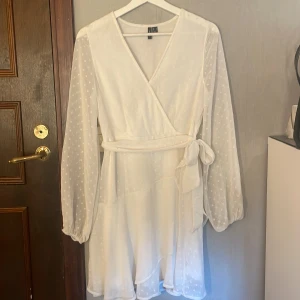 Vit omlottklänning med prickigt mönster - Superfin vit omlottklänning från Vera Moda med transparenta långärmar och diskret prickigt mönster. Klänningen har knyt i midjan, v-ringning och volangdetaljer nertill. Perfekt för sommarens alla tillfällen!