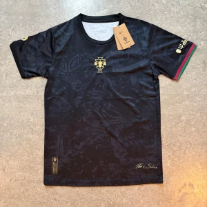 Portugal Nike svart fotbollströja - Svart Portugal fotbollströja från Nike med guldiga detaljer och nummer 7 på ryggen. Kortärmad modell i Dri-FIT-material med diskret mönster, broderat landslagsemblem och färgade ränder i grönt och rött på ena ärmen. Perfekt för dig som vill sticka ut på planen.