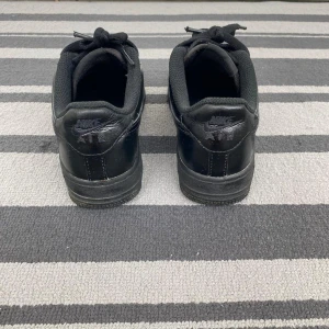 Svarta Nike Air Force 1 sneakers - Klassiska helsvarta Nike Air Force 1 sneakers i skinn med lågt skaft och rund tå. Skorna har svarta snören, perforerad tåbox och silverfärgad detalj med 'AF1' på snörningen. Perfekta för en clean och stilren look.