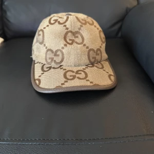 Beige keps från Gucci med GG-mönster - Snygg beige keps från Gucci med klassiskt GG-logomönster i brunt. Kepsen har justerbar rem baktill i brunt skinn och är tillverkad i bomullsmix. Perfekt för dig som vill sticka ut med en lyxig och tidlös accessoar.