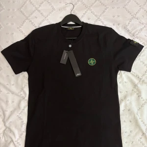 Stone Island t-shirt - Svart t-shirt från Stone Island med klassisk rund hals och ikonisk kompasspatch på bröstet och ärmen. Tillverkad i mjuk bomull för skön passform. Perfekt för dig som gillar streetwear och vill ha en clean look med tydlig logga.
