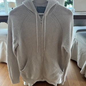 Beige stickad hoodie från Paul Taylor - Mysig beige stickad hoodie från Paul Taylor med dragkedja framtill och två stora fickor. Tröjan har huva med snörning och är tillverkad i en kashmirblandning som känns lyxig och mjuk. Perfekt för lager-på-lager och chill dagar.