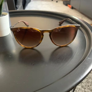Bruna Ray-Ban solglasögon - Snygga solglasögon från Ray-Ban med brunmelerad båge och klassisk form. Tunt metallskalmar och Ray-Ban-logga på glaset. Perfekta för att lyfta din stil i sommar.