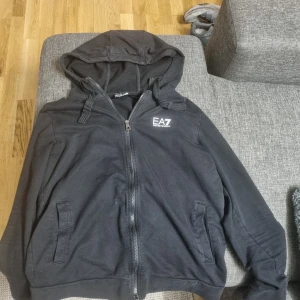Helt ny Svart hoodie från EA7 Emporio Armani - Svart hoodie från EA7 Emporio Armani med dragkedja och huva. Utan någon fel alls. Tröjan har två fickor framtill, snörning i huvan och en vit EA7-logga på bröstet. Tillverkad i mjuk bomull, perfekt för en chill och stilren look.
