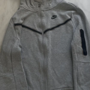 Grå zip-hoodie från Nike 158-170cm - Grå hoodie från Nike i storlek 158-170 cm med hel dragkedja, svart Nike-logga på bröstet och svarta detaljer över bröstet. Jackan har huva, lång ärm och en praktisk ficka med dragkedja på ena ärmen. Perfekt för chill eller träning men även sommarkvällar och liknande.