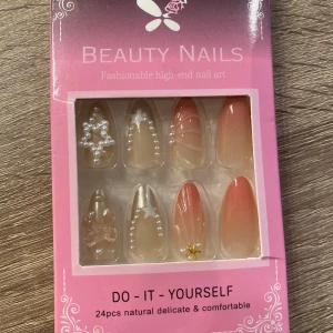 Press on-naglar med pärlor och stjärnor - Säljer ett set med 24 st press on-naglar i nude och rosa toner. Naglarna har mandelform och dekoreras med vita pärlor, stjärnor, guldstjärna och 3D-rosett. Inkluderar nagelfil och klisterlappar för enkel applicering. Perfekt för dig som vill ha snygga naglar snabbt!