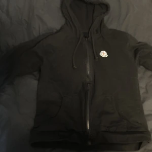 Svart hoodie från Moncler med dragkedja - Jag är 168 det står xl men det passar xs och s. Svart hoodie från Moncler med klassisk logga på bröstet. Tröjan har huva med snörning, hel dragkedja framtill och två fickor. Perfekt för dig som gillar en clean och stilren look. Passar både till jeans och mjukisbyxor.