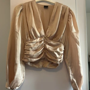 Blus - Superfin champagnefärgad blus från ginatricot med djup v-ringning, puffiga långärmar och snygg omlott-effekt. Framsidan har dekorativa knappar och rynkade detaljer som ger en lyxig känsla. Perfekt för dig som vill sticka ut lite extra.