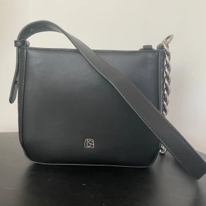 Svart axelväska från Lesac med kedjedetalj - Stilren svart axelväska från Lesac i slätt skinn med silverfärgad kedjedetalj på sidan. Väskan har minimalistisk design, justerbart axelband och diskret logga framtill. Perfekt för dig som gillar en clean look med en touch av metall.