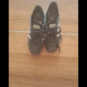 Svarta Adidas Spezial sneakers - Säljer ett par klassiska svarta Adidas spezial sneakers med vita ränder och snörning. Skorna har en gummisula med tydligt mönster och Adidas-logga. Perfekta för en sportig look. Mycket bra skick förutom lite missfärgning på bakre delen av tyget på vänster skon men inget som man tänker på.