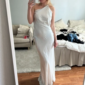 Vit axelbandslös långklänning - Elegant vit långklänning med en oneshoulder. Klänningen har en öppen rygg och är perfekt för speciella tillfällen. Den faller väldigt fint på kroppen och den öppna ryggen är inte heller allt för låg.