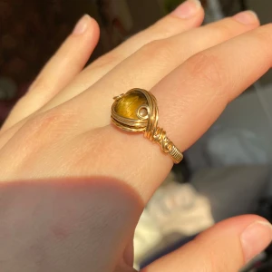 Guldfärgad ring med tigeröga - Snygg guldfärgad ring med en stor tigeröga-sten i mitten. Ringen har en unik design med detaljerade trådar som omsluter stenen. Perfekt för att ge en touch av elegans och mystik till din stil. - Nickelfri      - Non-tarnish behandling - Storleksguide på sista bilden
