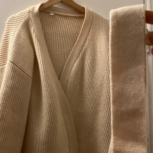 Beige ribbad kofta - Säljer en stilren beige ribbad kofta med omlott-design. Den har en mjuk och bekväm passform, perfekt för kyligare dagar. Koftan är långärmad och har ett bälte för att justera passformen. Perfekt för att hålla sig varm och snygg!