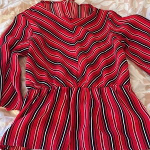Röd randig blus från Lindex - Säljer en färgglad röd blus från Lindex med svarta och vita ränder. Blusen har långa ärmar och en bekväm passform. Perfekt för att liva upp garderoben med lite färg!