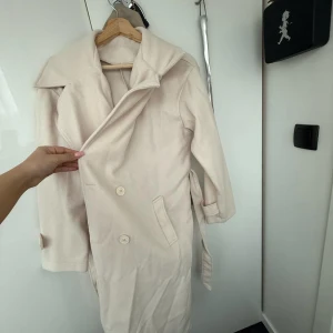Beige kappa med bälte - Säljer en stilren beige kappa med bälte och stora slag. Den har långa ärmar med dekorativa knappar vid ärmsluten och knäppning framtill. Perfekt för kyligare dagar!