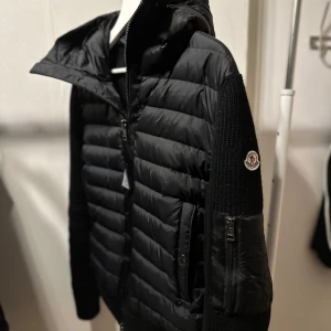 Svart pufferjacka från Moncler - Säljer en snygg svart pufferjacka från Moncler med stickade ärmar och dragkedja. Jackan har en quiltad design och en praktisk huva. Perfekt för kyligare dagar!