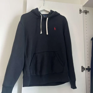 Svart hoodie från Polo Ralph Lauren - Säljer en klassisk svart hoodie från Polo Ralph Lauren med en röd logga på bröstet. Tröjan har en känguruficka och justerbar huva med vita snören. Perfekt för en avslappnad stil.
