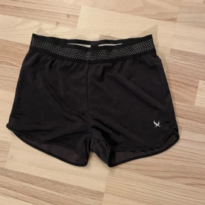 Svarta träningsshorts från SOC - Säljer ett par svarta träningsshorts från SOC med en bekväm elastisk midja och ventilerande material. Perfekta för intensiva träningspass eller löprundor. Shortsen har en stilren design med en liten logga på benet.