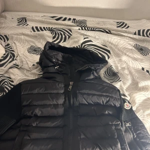 Svart pufferjacka från Moncler - Snygg svart pufferjacka från Moncler med stickade ärmar och dragkedja framtill. Jackan har en huva med snörning och en praktisk ficka med dragkedja på ärmen. Perfekt för kyliga dagar.