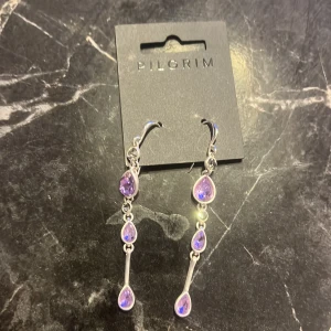 Lila örhängen från Pilgrim - Snygga och eleganta örhängen från Pilgrim med lila stenar i olika storlekar. De har en droppformad design och är perfekta för att ge en färgklick till din outfit. Passar både till vardag och festliga tillfällen.