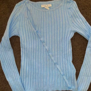 Ljusblå ribbad tröja  - Säljer en ljusblå ribbad tröja med en unik omlott-design och knappar framtill. Tröjan har långa ärmar och en stilren look som passar perfekt för både vardag och speciella tillfällen.