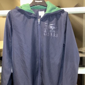 Mörkblå tracksuit från Lacoste - Säljer över delen av tracksuit/jacka från Lacoste med grön huva och dragkedja framtill. Jackan har en broderad logga och text på bröstet. Storlek 164 kan även passa xs. Väldigt bra skick inga tecken på användning 