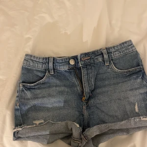 Blå jeansshorts med slitningar - Säljer ett par snygga blå jeansshorts med slitningar och upprullade kanter. De har en klassisk femficksdesign och stängs med dragkedja och knapp. Perfekta för en avslappnad stil. De är low waisted.