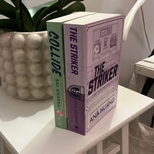 The Striker och Collide - Två böcker till salu: 'The Striker' av Ana Huang med ett sportigt omslag i lila och 'Collide' av Bal Khakra med ett mintgrönt omslag. Perfekt för dig som älskar romantik och sportteman. Böckerna står på ett vitt bord bredvid en dekorativ växt.