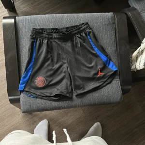 PSG dam shorts  - Snygga svarta träningsshorts från Jordan med blå detaljer och PSG-logga. Shortsen har en elastisk midja och praktiska fickor med dragkedjs fickor, Perfekta för sport och träning.