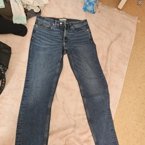 Blå jeansbyxor med normal passform - Säljer ett par klassiska blå jeansbyxor med normal passform. De har en midwaist design och är tillverkade i jeansmaterial. Byxorna har en dragkedja och knappar framtill samt fem fickor. Perfekta för en avslappnad stil.