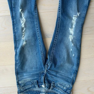 Blå bootcut jeans med slitningar - Snygga blå bootcut jeans med slitningar på framsidan. De har en klassisk femficksdesign och knappar på bakfickorna. 