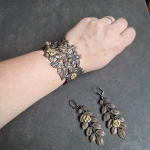Blomformade smycken i tenn - Säljer ett set med ett armband och örhängen i metall med vackra blommotiv. Armbandet har ett intrikat blommönster med guldfärgade detaljer, medan örhängena har hängande löv och en guldfärgad blomma. Perfekt för att ge en elegant touch till din outfit.