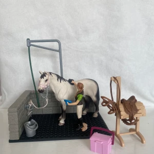 Hästvårdsset med häst, spolspilta och tillbehör  - Detaljerat hästvårdsset med en häst, människa och tillbehör. Inkluderar en tvättstation med hink och slang, en sadel och träns på sadelbock och en rosa ryktlåda med borstar. Perfekt för hästälskare som vill skapa realistiska stallscener.