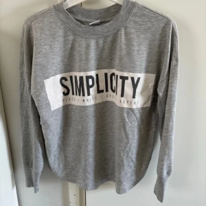 Grå tröja från Gina Tricot med text - Säljer en grå långärmad tröja från Gina Tricot med texten 'SIMPLICITY' i svart på bröstet. Tröjan har en rund halsringning och är perfekt för en avslappnad stil. Materialet är mjukt och bekvämt. Nopprig men går att få bort med noppmaskin